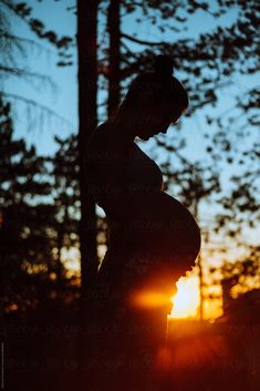 the silhouette of a pregnant woman in front of trees with the sun setting behind her