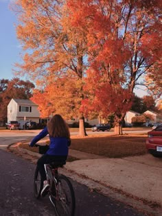 a woman riding her bike down the street in front of some trees with orange leaves