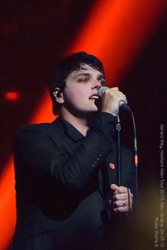 a young man singing into a microphone on stage at a music concert with red and black lighting behind him