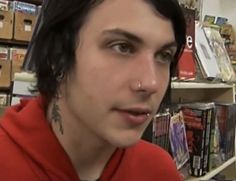 a man with black hair wearing a red hoodie in front of bookshelves