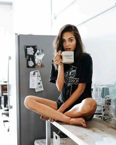 a woman sitting on top of a counter holding a coffee mug in her hand and looking at the camera