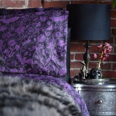 a bed with purple sheets and pillows in front of a brick wall next to a lamp