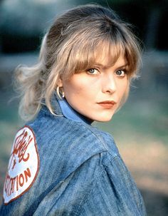 a woman with blonde hair wearing a denim jacket and earring, looking directly into the camera