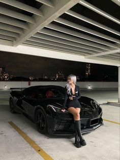 a woman sitting on the hood of a black sports car in a parking lot at night