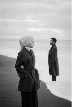 black and white photograph of two people standing on the beach looking at the ocean with one person in the distance