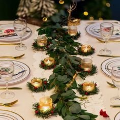 the table is set for christmas dinner with candles and greenery on it, along with other place settings