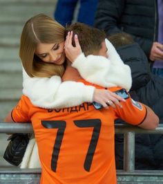 two people hugging each other in front of a crowd at a soccer game, one is wearing an orange jersey and the other has a white fur collar