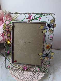 a decorative glass frame with flowers and dragonflies on the front is sitting on a lace doily next to a pink flower