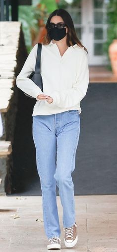 a woman in white sweater and jeans with black mask on her face walking down the street