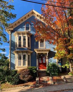 a blue two story house with red door and trees in the front yard on a fall day