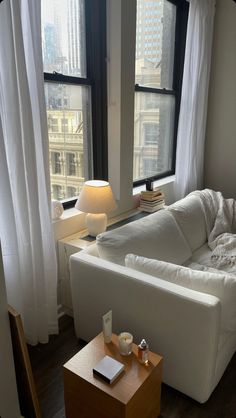 a white couch sitting in front of a window next to a table with a lamp on it