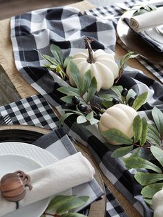 the table is set with white pumpkins and greenery on top of black checkered napkins