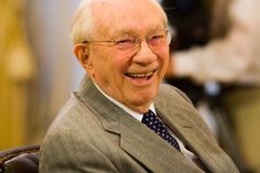 an older man in a suit and tie smiling at the camera while sitting on a chair