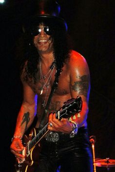 a shirtless man with long hair holding a guitar in his right hand and smiling