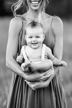 a woman holding a baby in her arms and smiling at the camera while standing in a field