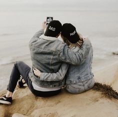 two people sitting on top of a sandy beach next to the ocean taking a selfie