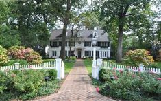 a large white house surrounded by trees and flowers on a brick walkway leading to the front door