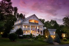 a large white house with lights on in the windows and trees around it at dusk