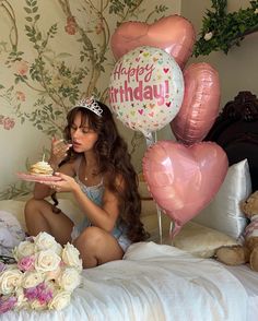 a woman sitting on a bed holding a piece of cake with balloons and flowers in the background