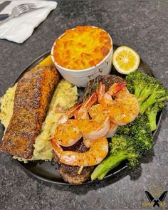 a black plate topped with shrimp, broccoli and grits next to a bowl of sauce