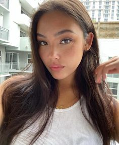 a woman with long brown hair wearing a white tank top and gold necklace looking at the camera