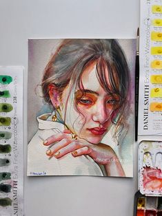 a painting of a woman's face with her hands on her chin, surrounded by watercolor paints