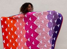 a woman holding up a colorful quilt on top of a white wall with an orange, pink, and purple design