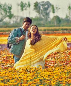 a man and woman standing in a field full of flowers holding a yellow scarf over their shoulders