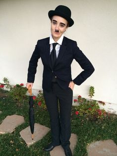 a man in a suit and hat holding an umbrella standing on the grass with his hands on his hips