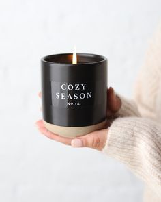 a woman holding a black candle in her hand with the words cozy season printed on it