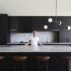 a man standing at the center of a kitchen island in front of black cabinets and bar stools