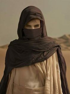 a woman wearing a black scarf and head covering in the desert with sand dunes behind her