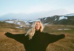 a woman standing in the middle of a field with her arms spread out and snow covered mountains behind her