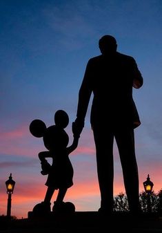 the silhouette of a man holding hands with a statue of mickey mouse in front of a sunset