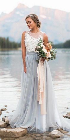 a woman standing on rocks in front of water wearing a gray and white wedding dress