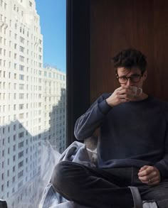 a man sitting in front of a window with his hand on his mouth while looking out the window