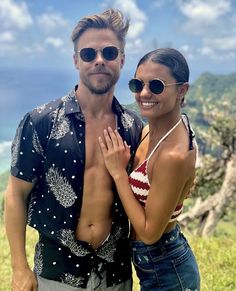 a man and woman standing next to each other on top of a hill with the ocean in the background