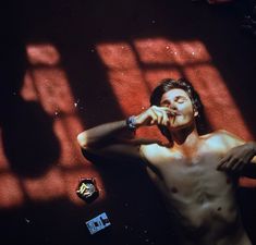 a shirtless man standing in front of a red wall holding his hand to his face