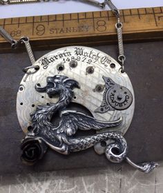 a silver necklace with a bird and moon design on it's side, sitting next to a ruler