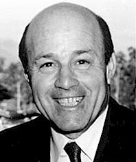 a black and white photo of a man in a suit smiling at the camera with trees in the background