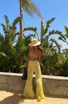 a woman in yellow pants and a hat is standing by some palm trees with her hand on her hip