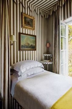 a bedroom with striped walls and white bedding in front of an open window that looks out onto the outdoors