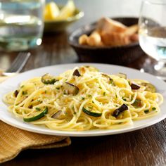 a white plate topped with pasta and vegetables on top of a wooden table next to wine glasses