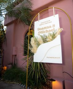 a sign hanging from the side of a building next to a palm tree and potted plant