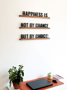 a desk with a laptop, potted plant and two wooden shelves on the wall that say happiness is not by chance but by choice