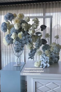 a vase filled with white and blue flowers on top of a table next to a window