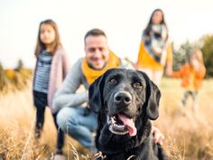 a man kneeling down next to a black dog in a field with two children behind him