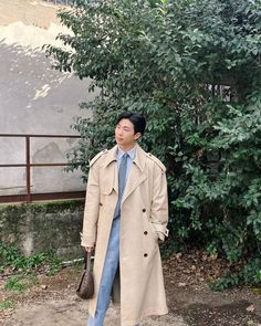 a man in a trench coat and tie is standing by a tree with his hand on his hip