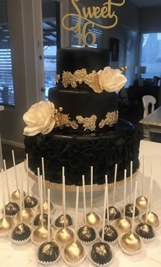 there is a black and gold cake with white flowers on the top, surrounded by chocolate lollipops