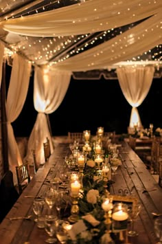 a long wooden table topped with lots of candles next to white drapes and lights